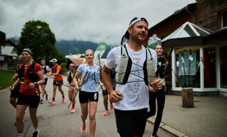 A group of runners is jogging along a road. The sky is overcast, and the surroundings appear mountainous and green. | © Kleinwalsertal Tourismus | Oliver Farys