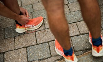 A man ties his sports shoes with an orange sole. The surface consists of gray cobblestones. | © Kleinwalsertal Tourismus | Oliver Farys