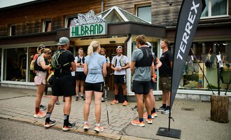 A group of runners gathers in front of a sports store. In the background, a sign and a promotional flag can be seen. | © Kleinwalsertal Tourismus | Oliver Farys