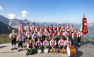 A large music band in traditional costumes stands on a wooden platform with a mountain view. Beautiful alpine landscapes are visible in the background. | © Trachtenkapelle Riezlern