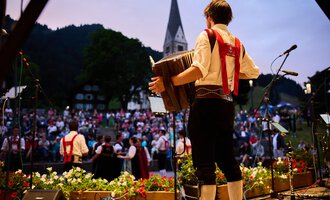 A musician plays an accordion in front of a large crowd. In the background, a church and a picturesque landscape can be seen. | © Kleinwalsertal Tourismus | Oliver Farys