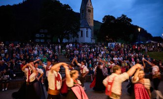 A crowd gathers in front of a church as dancers in traditional costumes perform. The atmosphere is festive and lively at dusk. | © Kleinwalsertal Tourismus | Oliver Farys