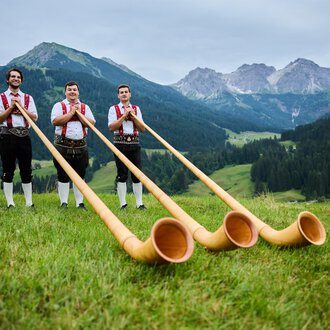 © Kleinwalsertal Tourismus eGen | Oliver Farys Three men in traditional attire stand in a green meadow with an impressive mountain landscape in the background. They hold large alpine horns in their hands. | © Kleinwalsertal Tourismus eGen | Oliver Farys