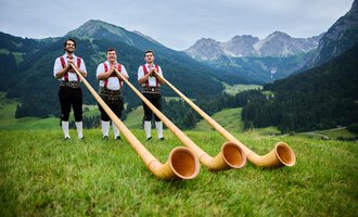 Drei Männer in traditioneller Tracht stehen in einer grünen Wiese mit einer beeindruckenden Berglandschaft im Hintergrund. Sie halten große Alphörner in den Händen. | © Kleinwalsertal Tourismus eGen | Oliver Farys