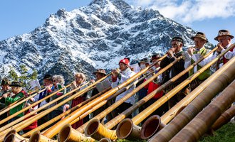 Eine Gruppe von Musikern spielt Alphorn in den Bergen. Im Hintergrund sind schneebedeckte Gipfel und ein blauer Himmel zu sehen. | © Kleinwalsertal Tourismus eGen | Frank Drechsel