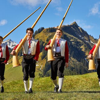 Vier Männer in traditionellen Trachten tragen große Alphörner. Im Hintergrund sind grüne Wiesen und beeindruckende Berge zu sehen. | © Kleinwalsertal Tourismus | Frank Drechsel