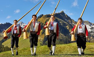 Vier Männer in traditionellen Trachten tragen große Alphörner. Im Hintergrund sind grüne Wiesen und beeindruckende Berge zu sehen. | © Kleinwalsertal Tourismus | Frank Drechsel