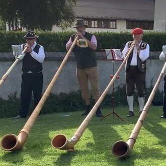 © Alphornfreunde grenzenlos A group of five men is playing alphorns on a meadow. They are wearing traditional costumes and are standing in a picturesque setting. | © Alphornfreunde grenzenlos