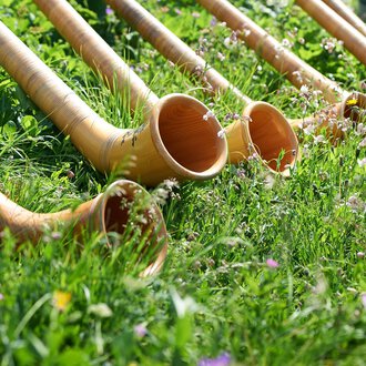 © OBERSTDORF · KLEINWALSERTAL BERGBAHNEN A series of alphorns lies in the grass. The horns are made of wood and surrounded by colorful flowers. | © OBERSTDORF · KLEINWALSERTAL BERGBAHNEN