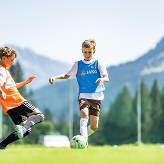 © Fußballschule Anstoß Young soccer players on a green field with mountains in the background. One player is dribbling the ball while another is trying to stop him. | © Fußballschule Anstoß