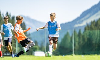 Young soccer players on a green field with mountains in the background. One player is dribbling the ball while another is trying to stop him. | © Fußballschule Anstoß