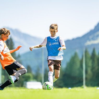 Young soccer players on a green field with mountains in the background. One player is dribbling the ball while another is trying to stop him. | © Fußballschule Anstoß