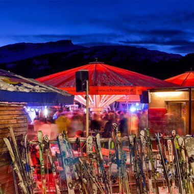 © Plattform Kleinwalsertal | Michael Monschau A ski lodge with red sun umbrellas in a wintry setting. In the foreground, several skis are visible, and in the background, people can be seen enjoying the atmosphere. | © Plattform Kleinwalsertal | Michael Monschau