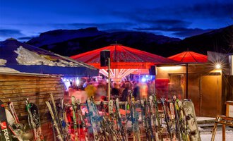 © Plattform Kleinwalsertal | Michael Monschau A ski lodge with red sun umbrellas in a wintry setting. In the foreground, several skis are visible, and in the background, people can be seen enjoying the atmosphere. | © Plattform Kleinwalsertal | Michael Monschau