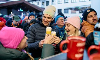 © Kleinwalsertal Tourismus | Oliver Farys A cheerful group of people toasts with drinks. They are wearing warm jackets and hats and are standing in a lively crowd. | © Kleinwalsertal Tourismus | Oliver Farys