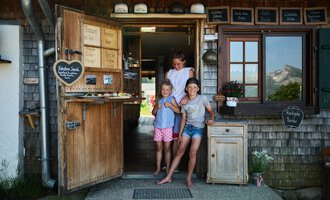 An inviting entrance door to a small shop, surrounded by wood and greenery. Three children are happily standing in front of it, smiling at the camera. | © Kleinwalsertal Tourismus | Oliver Farys