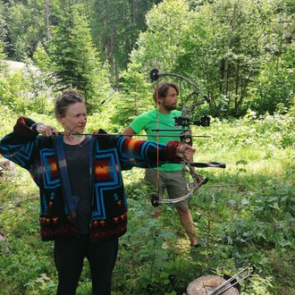 © Wildnisschule Kleinwalsertal | Ronny Katz Two people in the woods shooting arrows. The woman is wearing a colorful sweater and aims with a bow, while the man stands behind her in a green T-shirt. | © Wildnisschule Kleinwalsertal | Ronny Katz