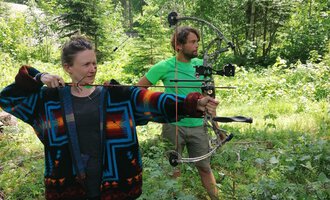 Two people in the woods shooting arrows. The woman is wearing a colorful sweater and aims with a bow, while the man stands behind her in a green T-shirt. | © Wildnisschule Kleinwalsertal | Ronny Katz