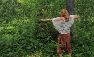 A woman stands in the forest aiming with a bow. Around her are lush green plants and trees.