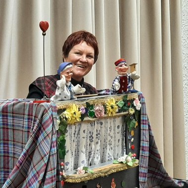 Eine Frau präsentiert ein Puppenspiel mit zwei handgefertigten Marionetten. Der Hintergrund ist mit einem hellen Vorhang und einer bunten Dekoration geschmückt. | © Marie Luise Kaiser | Veronika Senn