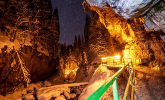 © Breitachklammverein eG | Dominik Berchtold A narrow, snow-covered path winds through a deeply snow-laden gorge. The night sky is dotted with stars, while warm lights illuminate the surroundings. | © Breitachklammverein eG | Dominik Berchtold