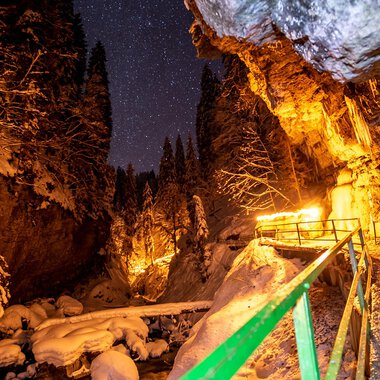 © Breitachklammverein eG | Dominik Berchtold A narrow, snow-covered path winds through a deeply snow-laden gorge. The night sky is dotted with stars, while warm lights illuminate the surroundings. | © Breitachklammverein eG | Dominik Berchtold