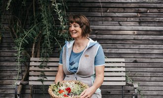 A smiling woman is sitting on a bench holding a basket of fresh plants. In the background, wooden slats and green plants can be seen. | © Naturhotel Chesa Valisa