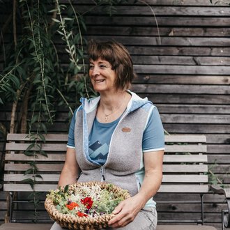 © Naturhotel Chesa Valisa A smiling woman is sitting on a bench holding a basket of fresh plants. In the background, wooden slats and green plants can be seen. | © Naturhotel Chesa Valisa