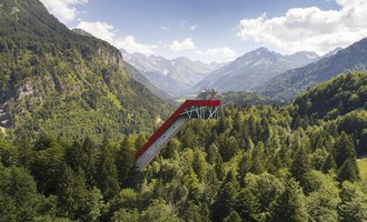 © Skiflugschanze Oberstdorf | Eren Karaman An impressive observation platform that extends above the trees, surrounded by majestic mountains and dense forests. The sky is clear and shows some clouds. | © Skiflugschanze Oberstdorf | Eren Karaman