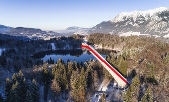 © Skiflugschanze Oberstdorf | Eren Karaman A long bridge with red and white stripes spans a tranquil, snow-covered valley and a lake. In the background, majestic mountains can be seen, glowing in the winter sun. | © Skiflugschanze Oberstdorf | Eren Karaman