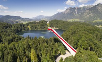 © Skiflugschanze Oberstdorf | Eren Karaman An impressive viewpoint bridge over a clear lake, surrounded by green forests and mountains. The sky is blue with some clouds. | © Skiflugschanze Oberstdorf | Eren Karaman