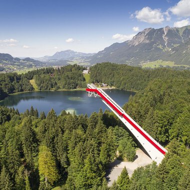 © Skiflugschanze Oberstdorf | Eren Karaman An impressive viewpoint bridge over a clear lake, surrounded by green forests and mountains. The sky is blue with some clouds. | © Skiflugschanze Oberstdorf | Eren Karaman