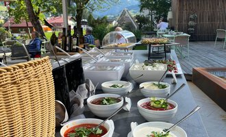 A beautifully set table outside with various bowls full of fresh salads and soups. In the background, there are trees and an inviting terrace. | © Genuss- und Aktivhotel Sonnenburg