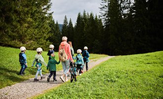 A group of children wearing helmets walks along a path, accompanied by an adult. The surroundings consist of green meadows and tall trees. | © Kleinwalsertal Tourismus | Oliver Farys