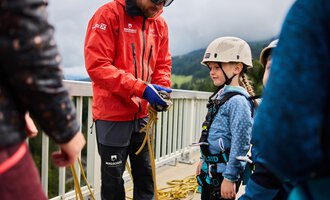 A trainer is explaining to a child the safe handling of climbing equipment. In the background, more children and a green mountain landscape can be seen. | © Kleinwalsertal Tourismus | Oliver Farys