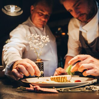 Two chefs working on a plate while carefully adding ingredients. The atmosphere is warm and inviting, with a focus on the artistic presentation of the dish. | © Hotel Birkenhöhe Bantel GmbH & Co KG