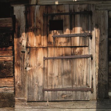 An old wooden plank door with iron fittings. The door has a small window and is surrounded by a rustic wall. | © R. Koeberle