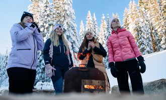 © Kleinwalsertal Tourismus | Oliver Farys A group of women stands around a small heater in a snowy winter landscape. In the background, snow-covered trees and a clear blue sky can be seen. | © Kleinwalsertal Tourismus | Oliver Farys