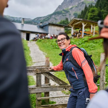 A cozy mountain house surrounded by green meadows and tall mountains. In the foreground, a path leads to the front door, and a flag is waving in the wind. | © Kleinwalsertal Tourismus eGen - Fotograf: Oliver Farys