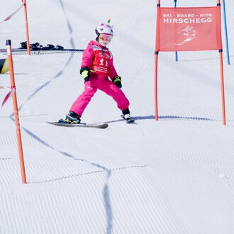 A small skier in pink ski gear is skiing through a gate on a snowy slope. In the background, other skiers and color markings can be seen. | © Skischule Hirschegg | Clemens Paul