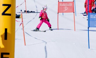 © Skischule Hirschegg | Clemens Paul A small skier in pink ski gear is skiing through a gate on a snowy slope. In the background, other skiers and color markings can be seen. | © Skischule Hirschegg | Clemens Paul