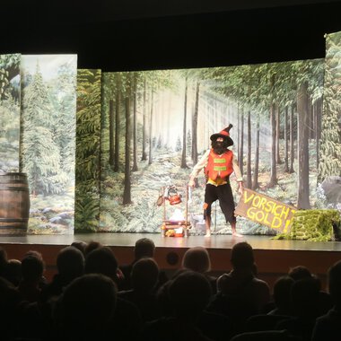 © KiWi Kindertheater GbR A theater performance with a colorful stage design that depicts a forest. An actor in a costume interacts with the audience. | © KiWi Kindertheater GbR