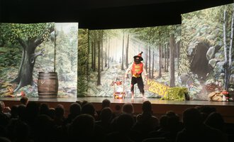 A theater performance with a colorful stage design that depicts a forest. An actor in a costume interacts with the audience. | © KiWi Kindertheater GbR