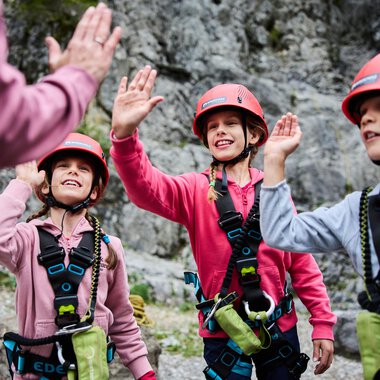 Three children in climbing gear greet an adult. They are wearing helmets and smiling against a rocky backdrop. | © Kleinwalsertal Tourismus | Oliver Farys
