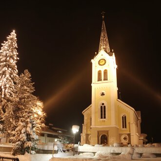 A snowy landscape with a illuminated church and a decorated Christmas tree. The night sky enhances the festive atmosphere. | © Kleinwalsertal Tourismus | Frank Drechsel