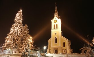 A snowy landscape with a illuminated church and a decorated Christmas tree. The night sky enhances the festive atmosphere. | © Kleinwalsertal Tourismus | Frank Drechsel