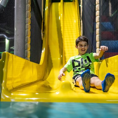 © Wonnemar Sonthofen | interSPA Gruppe A smiling child happily slides down a yellow slide. In the background, playground equipment can be seen. | © Wonnemar Sonthofen | interSPA Gruppe