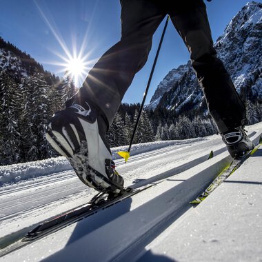 Ein Skifahrer gleitet über eine schneebedeckte Langlaufstrecke. Im Hintergrund leuchten die Berge unter einem klaren blauen Himmel. | © Kleinwalsertal Tourismus | Dominik Berchtold
