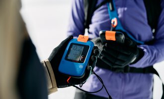 © Kleinwalsertal Tourismus | Oliver Farys A person is holding a transceiver device in their hand. In the background, a snowy area is visible. | © Kleinwalsertal Tourismus | Oliver Farys
