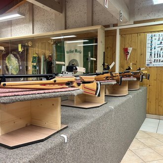 © Luftgewehrstand | Michael Veit A series of sporting rifles on display stands in a shooting range. In the background, instructions and information about shooting sports can be seen. | © Luftgewehrstand | Michael Veit